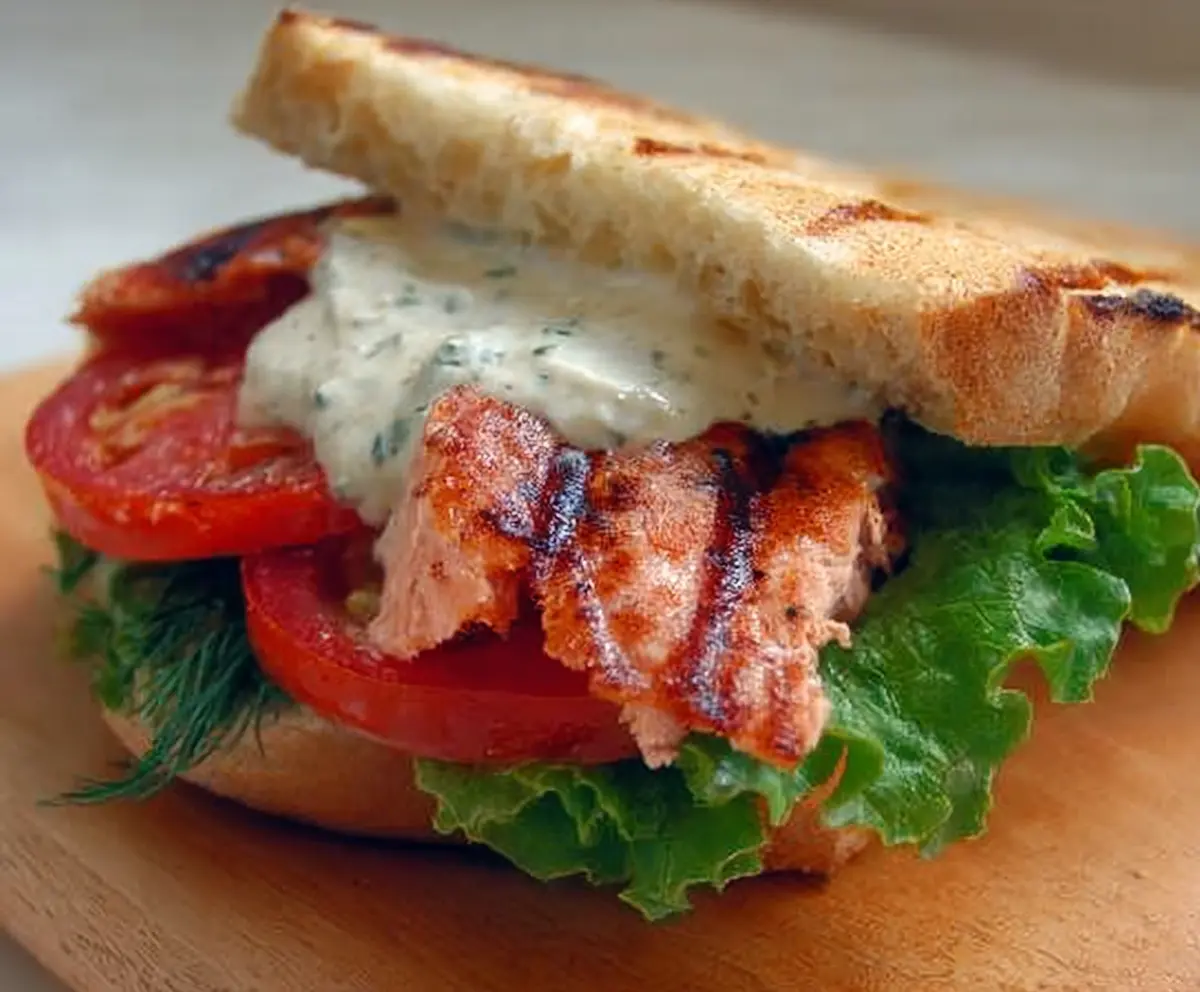 Delicious grilled salmon sandwich topped with fresh dill sauce on a rustic plate.
